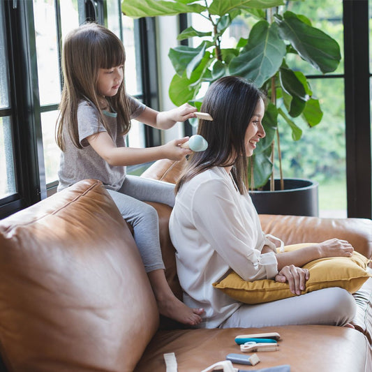 Child and mother playing with pretend hairdresser kit on a brown leather couch.