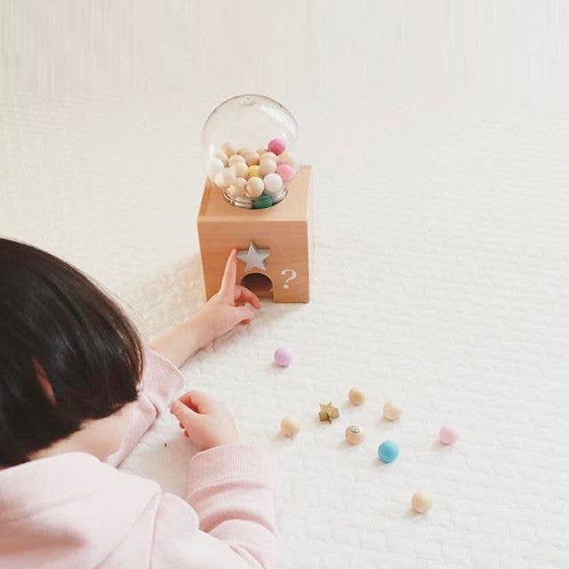 A child laying on the floor playing with a wooden toy resembling a gumball machine with a glass top filled with multicolored beads. Below the glass top, there's a star-shaped button and a question mark.