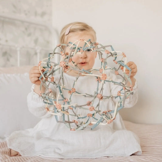 A child sitting on a bed holding a multicolored expandable Hoberman sphere, also known as the Mindfulness Magic Ball.