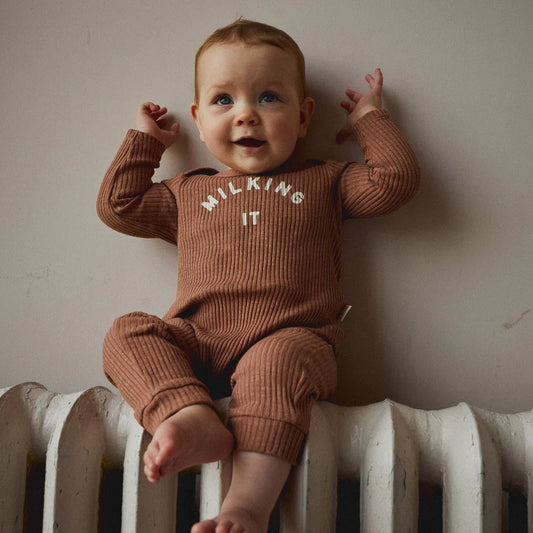 A baby wearing a brown onesie with the text 'MILKING IT' printed on the front, sitting on a heater.