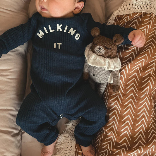 A navy blue onesie with the text 'MILKING IT' printed on the front, worn by a baby, accompanied by a brown teddy bear.