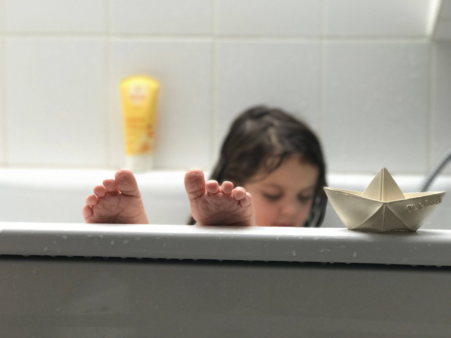 Child in bathtub with feet and origami boat on the edge of the tub.