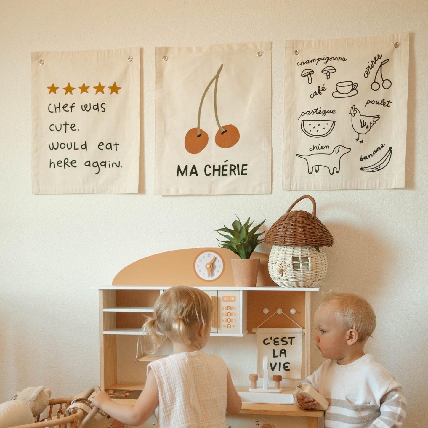 Children playing with a toy kitchen set in a room with decorative wall hangings.