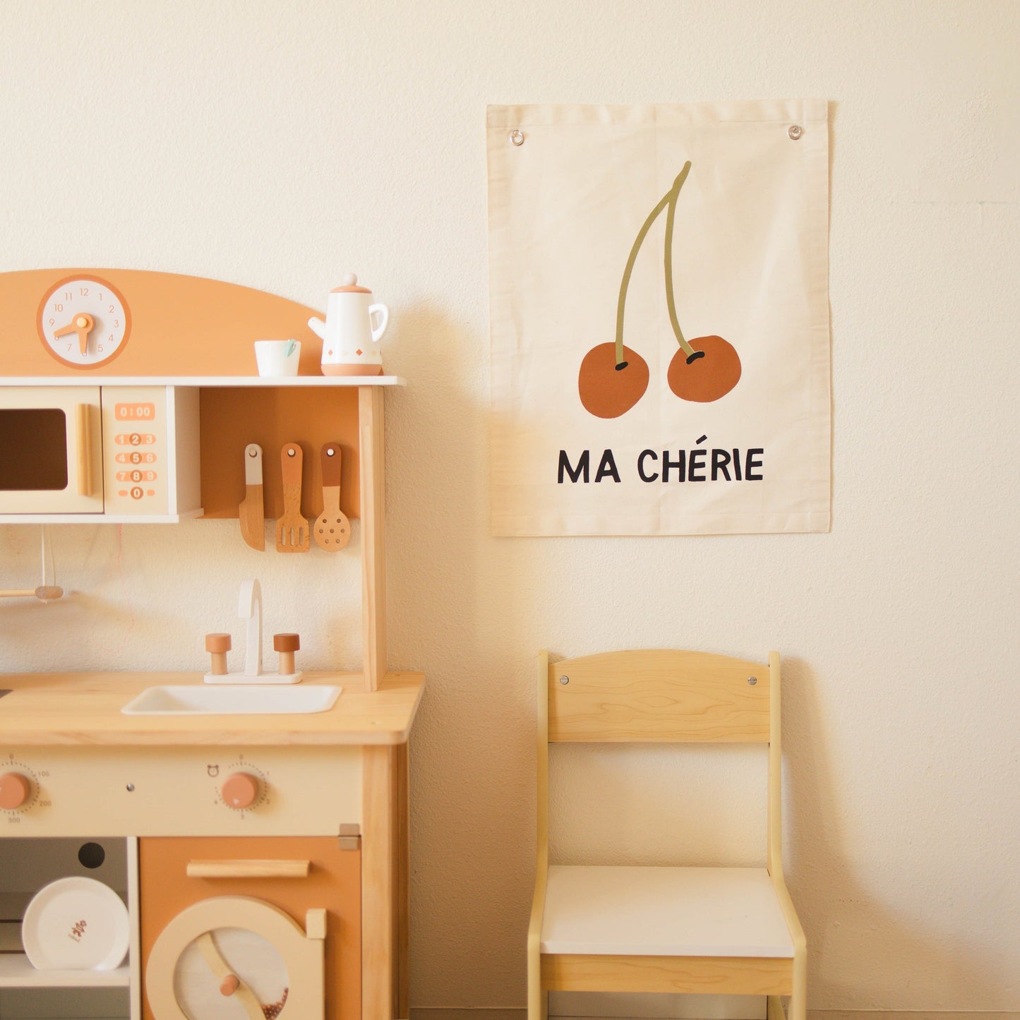Children's play kitchen set with a chair and decorative wall hanging on a light beige wall.