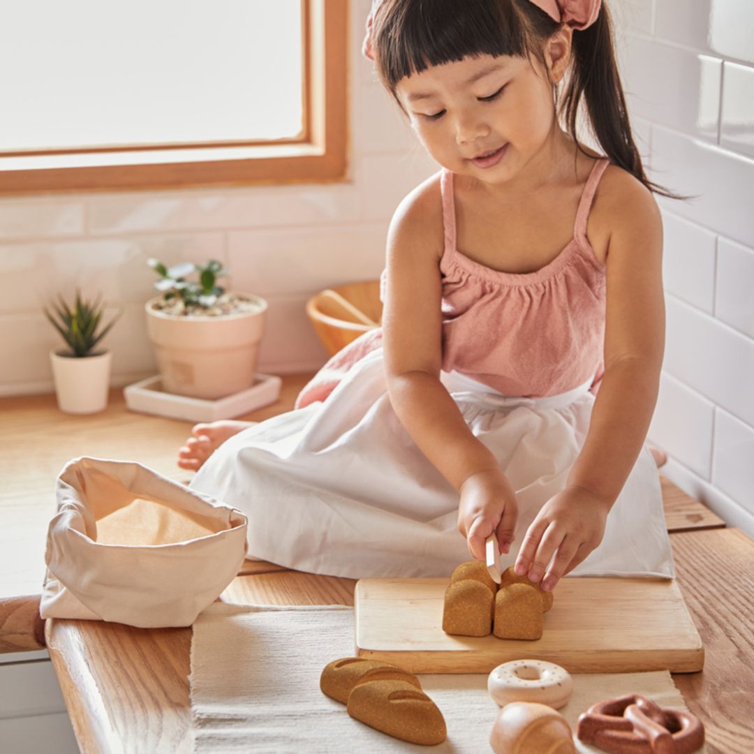Young girl playing with toy bread on a wooden cutting board in a kitchen.