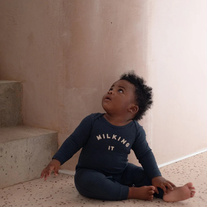 Baby boy sitting on the floor in front of steps wearing a navy blue onesie with the text 'MILKING IT' printed on the front.