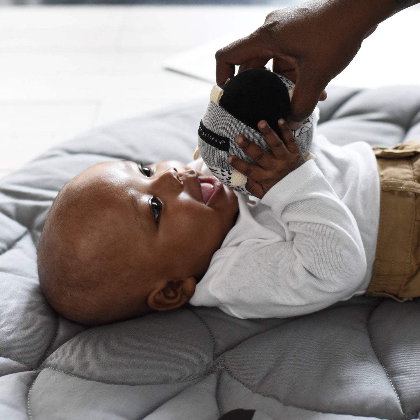A baby laying on a mat holding a sensory taggy ball with a woodlands theme, including animals and nature elements, with different textures and a rattle inside.