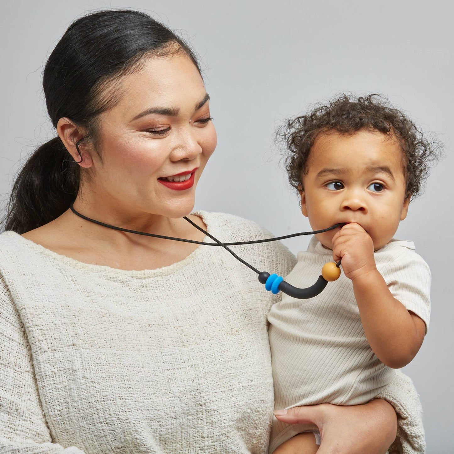 A woman wearing a teething necklace with a combination of solid colored beads in orange, black, and blue, connected by a black cord holding a baby who is chewing on the necklace.