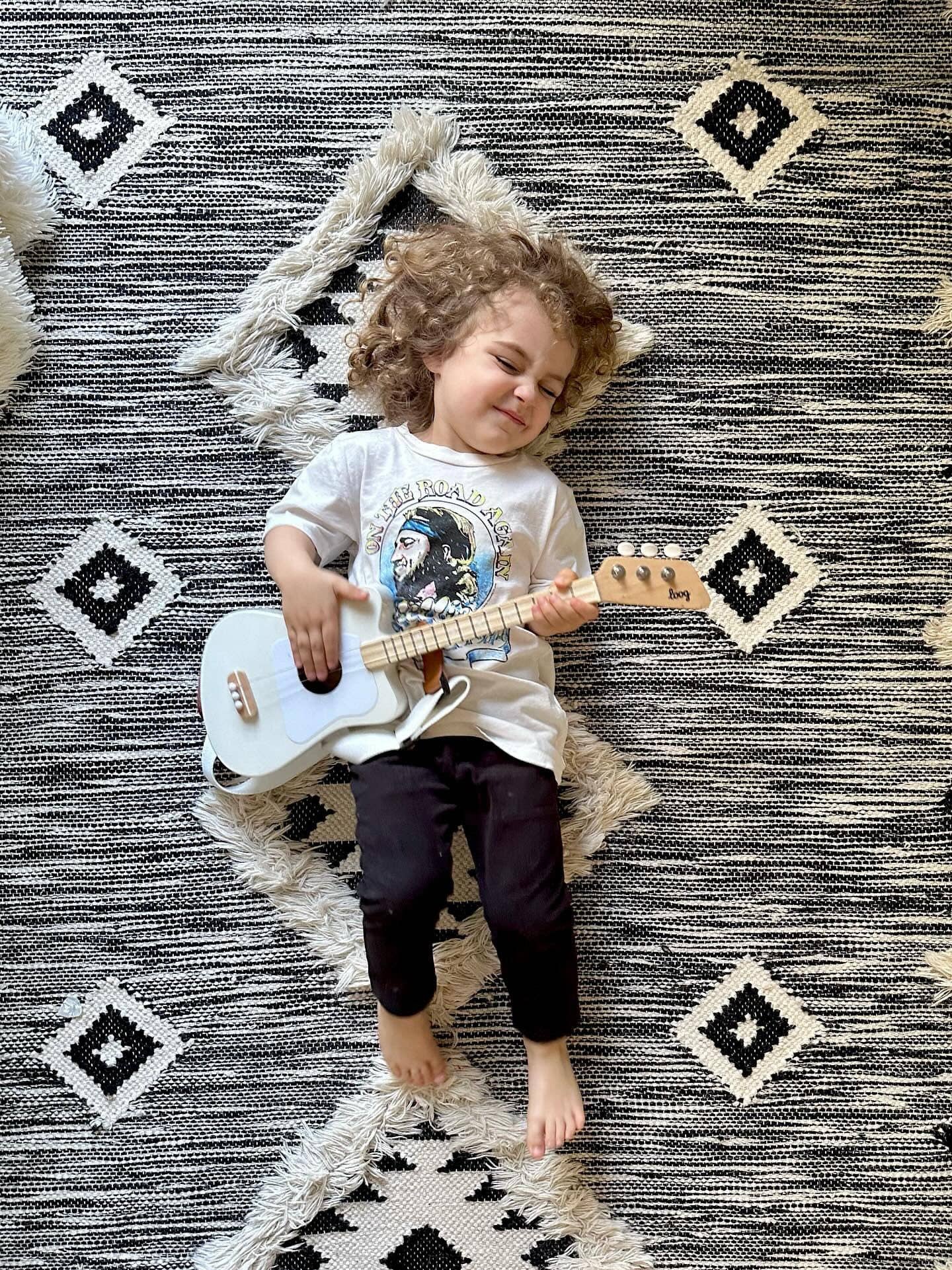 Toddler in Willie Nelson tee playing a Loog Mini Guitar laying on a carpet.