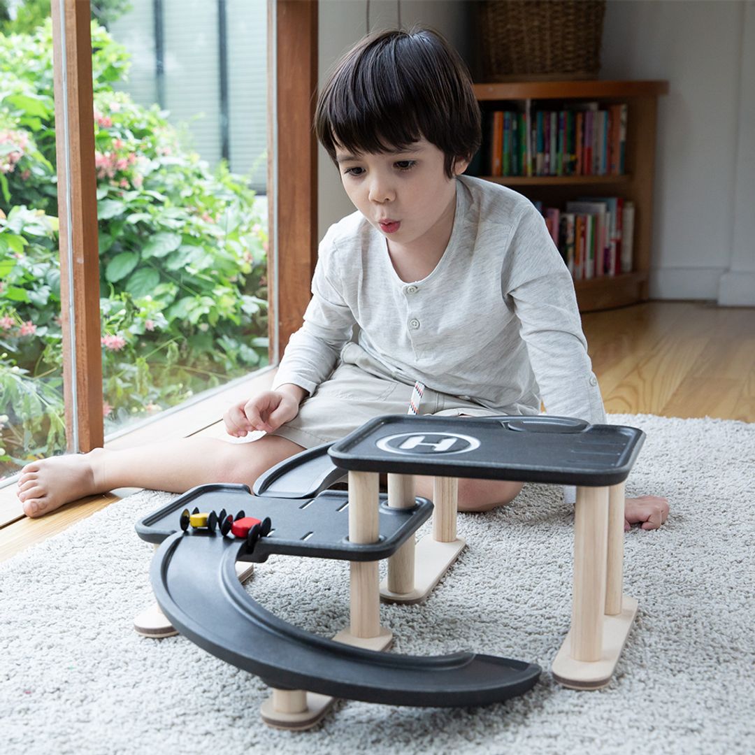 Child playing with a toy track set on a rug in a room with a bookshelf and window.