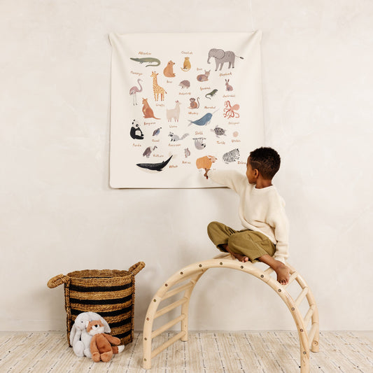 Child sitting on a wooden stool pointing at a wall chart with animal illustrations in a room.