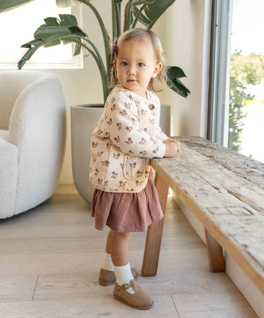 Toddler girl wearing a quilted v-neck button jacket with a 'fig flora' floral pattern on a beige background, featuring two front pockets and buttons down the front.