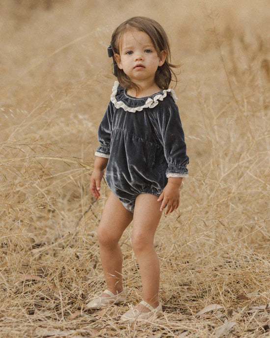 A toddler wearing a romper in a midnight hue, featuring a gathered bubbled body, legs, and sleeves with a ruffled collar and button backing. The fabric appears to be soft and textured.
