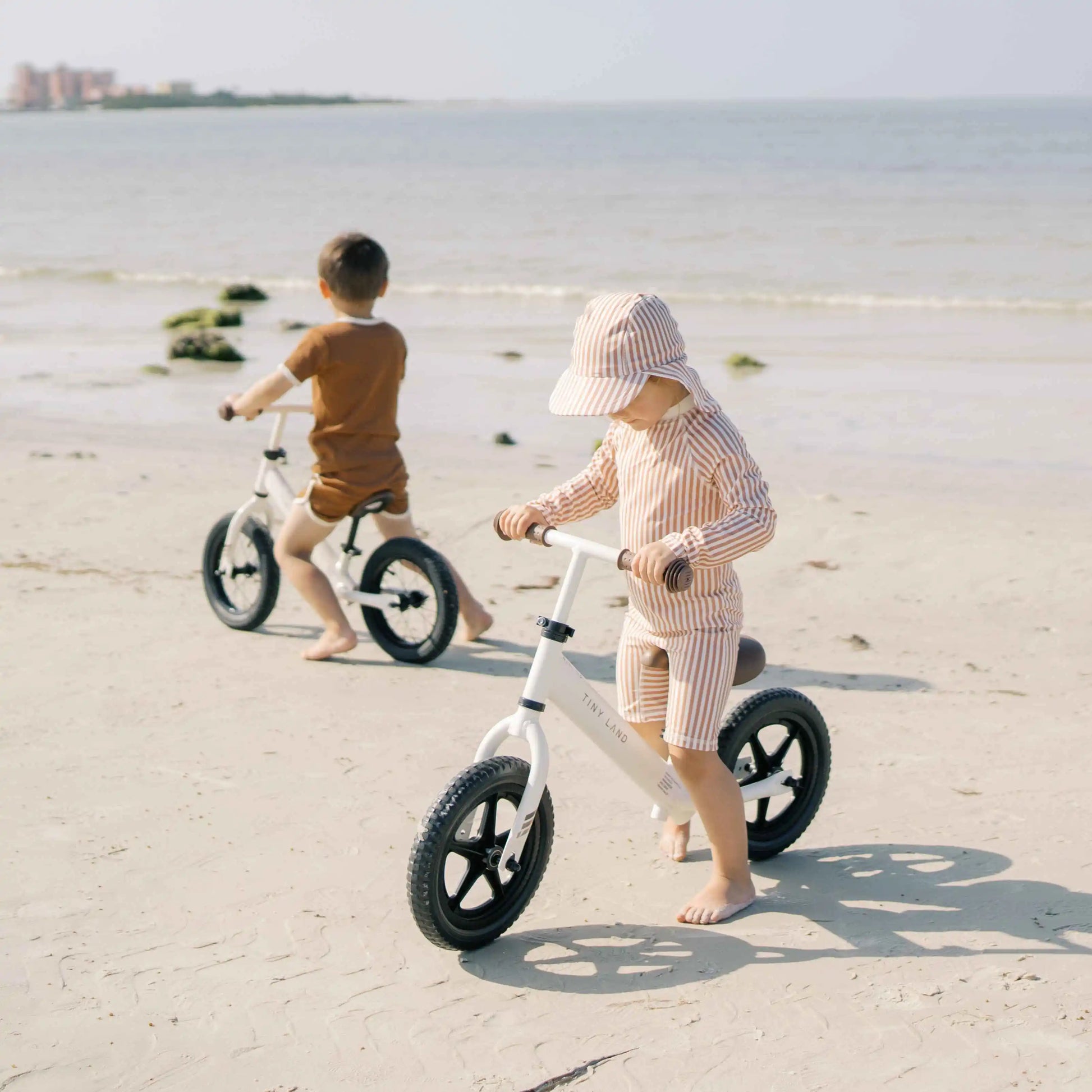 Two children riding balance bikes on a sandy beach.