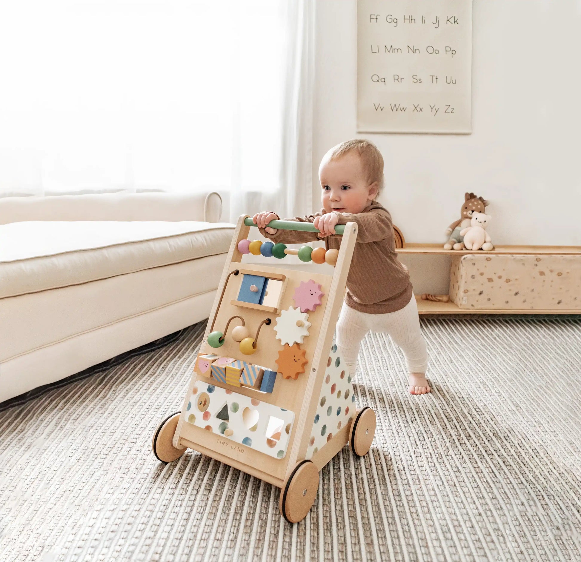 Baby playing with a wooden activity walker in a room with a white couch and shelves.