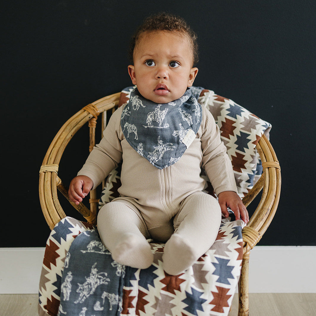 Baby sitting on a wicker chair with a patterned blanket, wearing a beige outfit and blue bib.