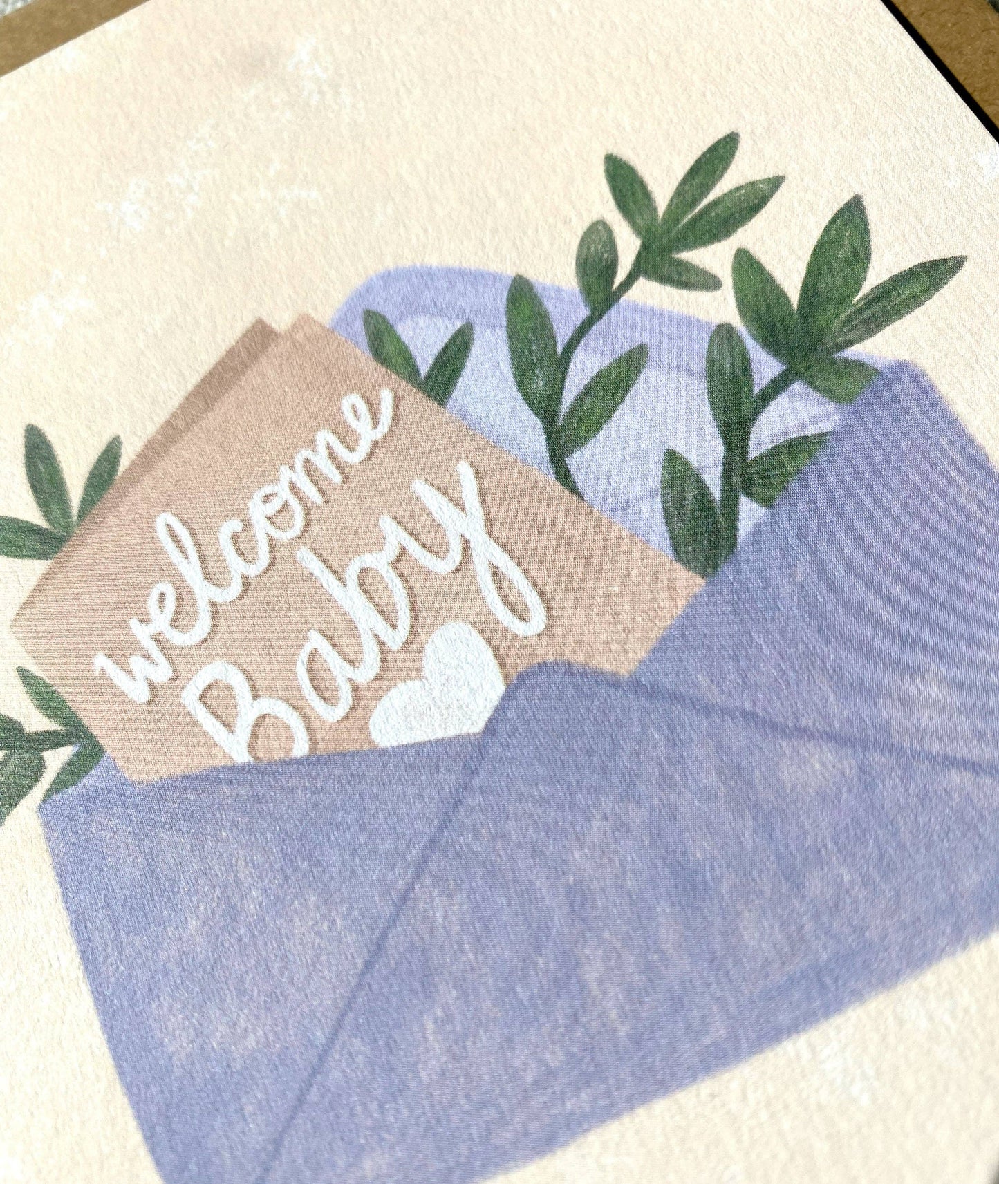 Close up of Welcome Baby card with the text 'Welcome Baby' and greenery coming out of a blue envelope.