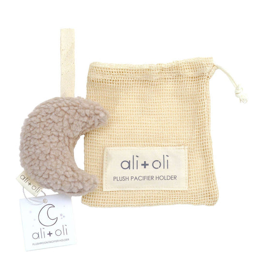 A beige-colored plush moon-shaped pacifier holder with a cardboard tag attached displaying 'ali+oli' branding, and a fabric tag attached to it as well.