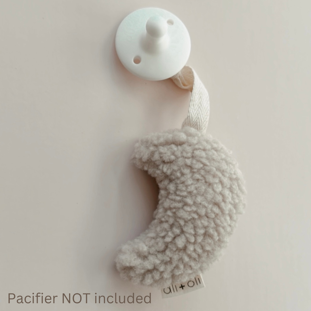 A white pacifier with a plush moon shaped pacifer holder on a cream background.