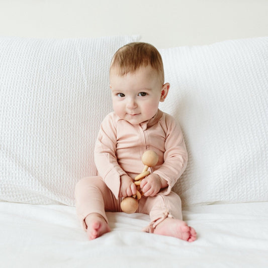 Baby in a pink outfit sitting on a white couch holding a wooden toy.