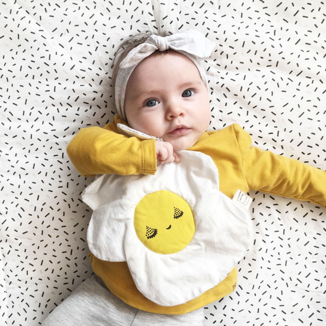 Baby wearing a yellow long-sleeve shirt and playing with a egg crinkle toy with a smiley face design on a patterned background.