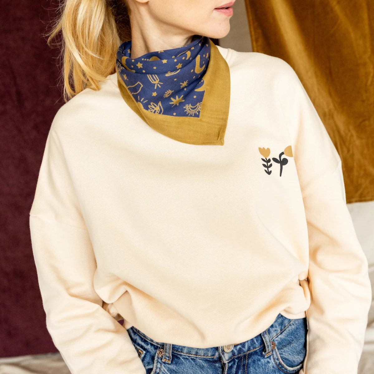 Woman wearing a beige sweater with a logo and a blue scarf.