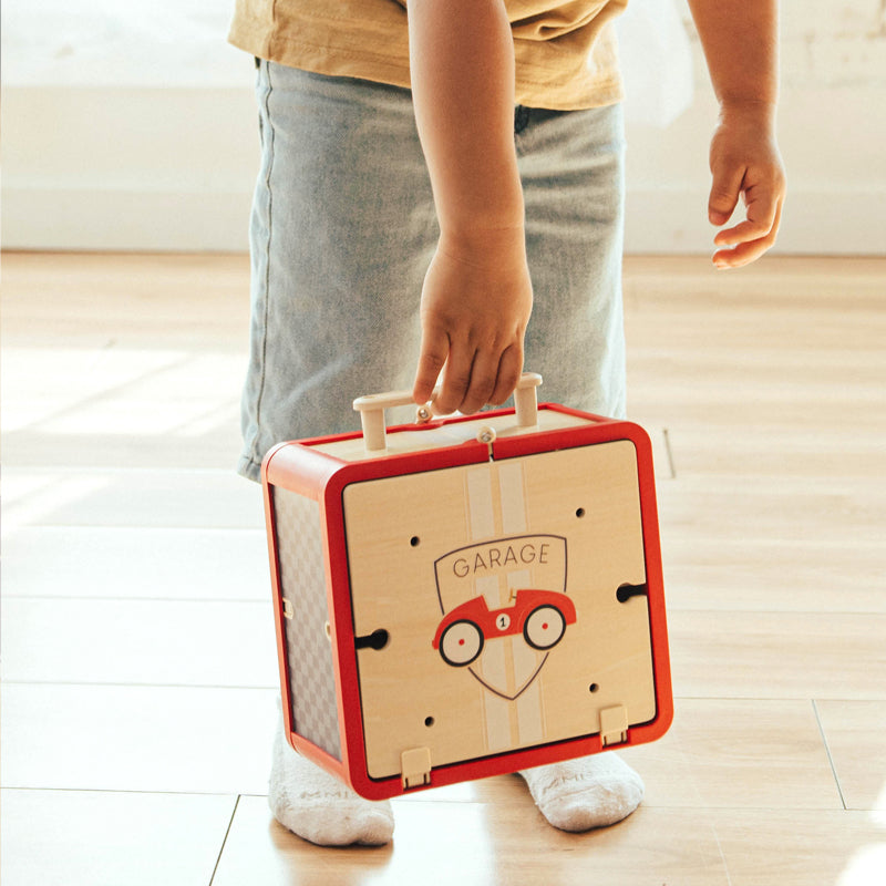 Child holding a red and beige toy suitcase with a car design on a wooden floor.