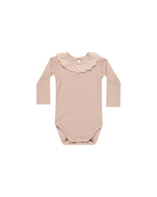 A blush-colored long sleeve bodysuit with a ruffled collar and snaps at the inseam.