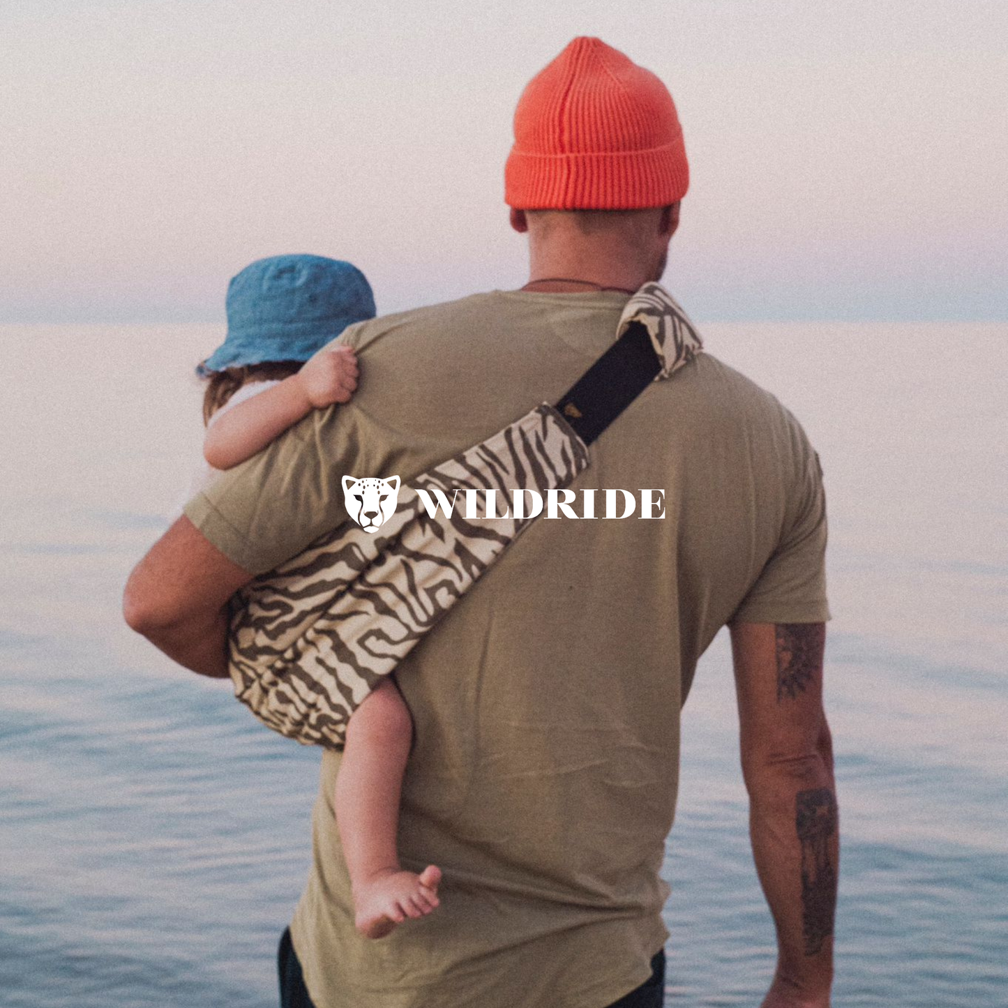 Man carrying a child on his back with a 'Wildride' bag, standing on a beach.