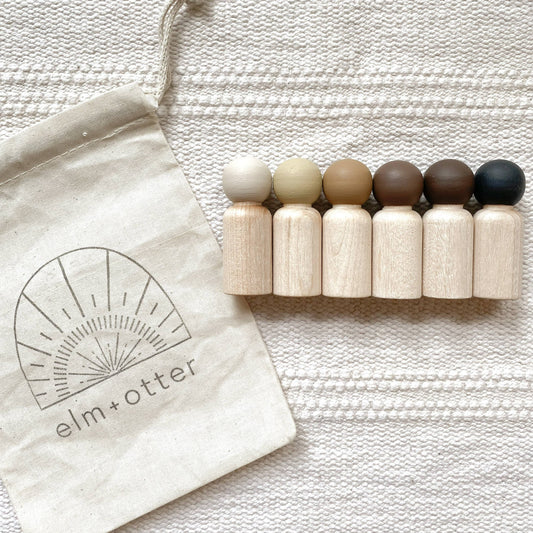 A set of 6 wooden peg dolls in various skin color shades with an Elm + Otter bag.