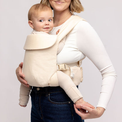 Woman holding a baby in a beige baby carrier against a plain background