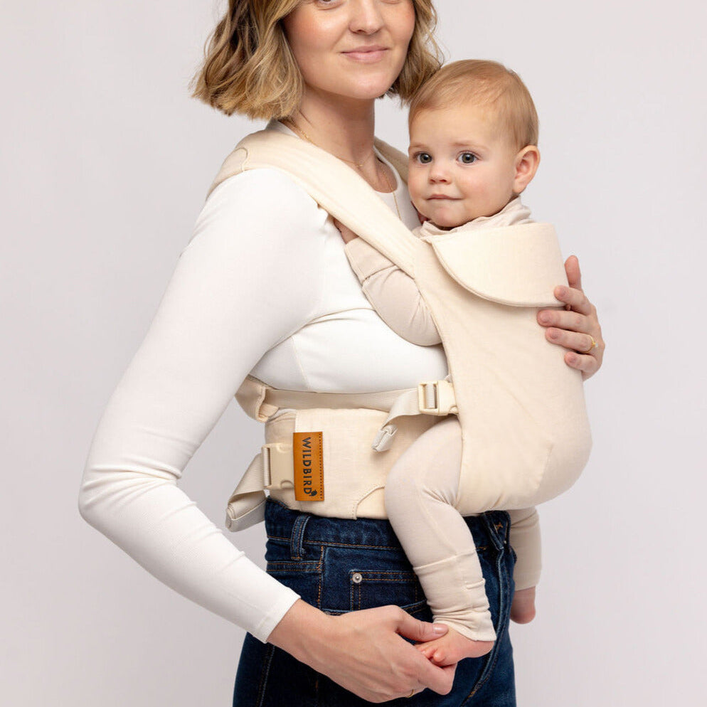 Woman holding a baby in a beige baby carrier against a plain background