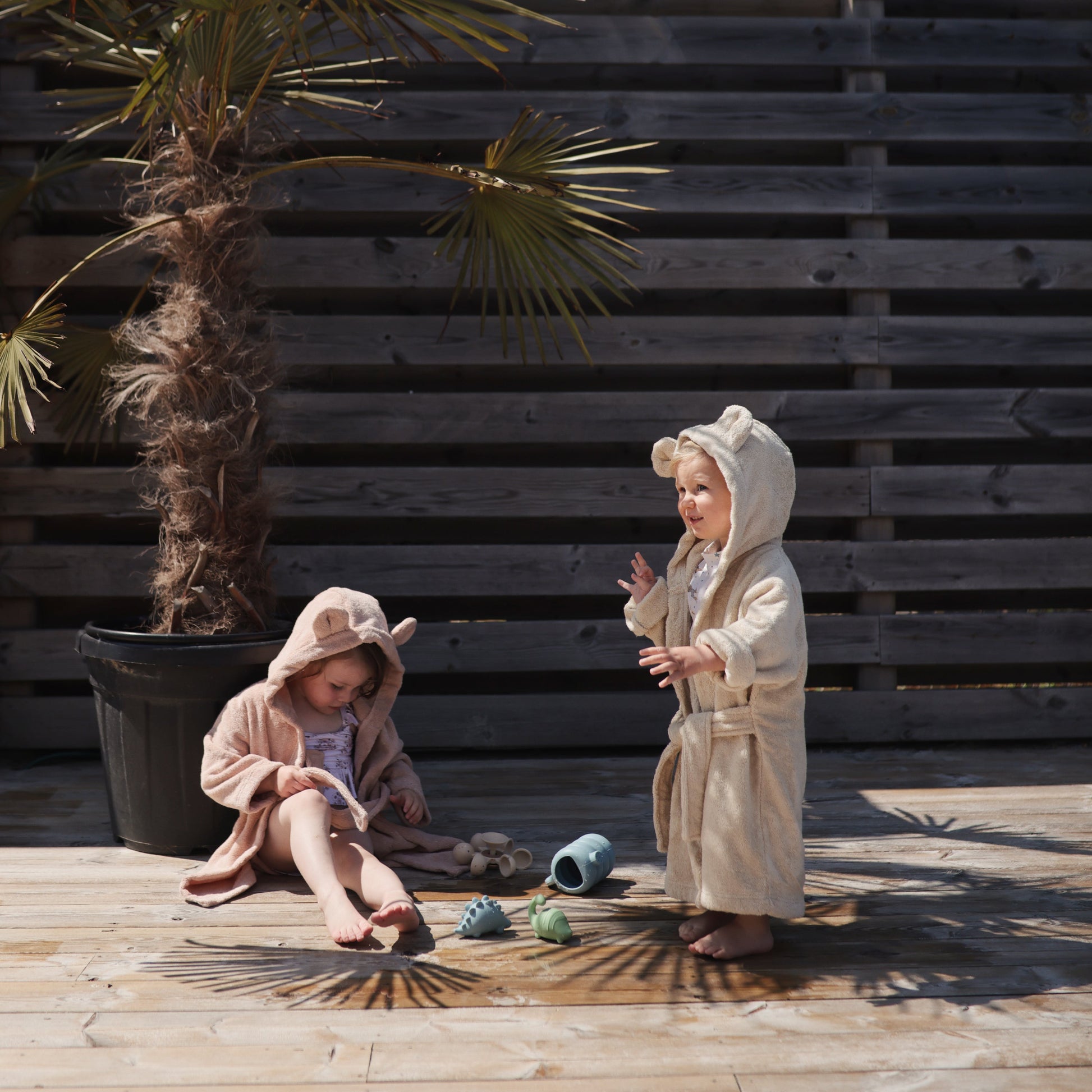 Two children in bathrobes standing on a wooden deck with a wooden wall and plant in the background.