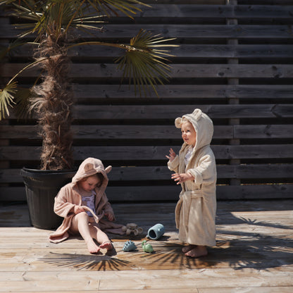 Two children in bathrobes standing on a wooden deck with a wooden wall and plant in the background.