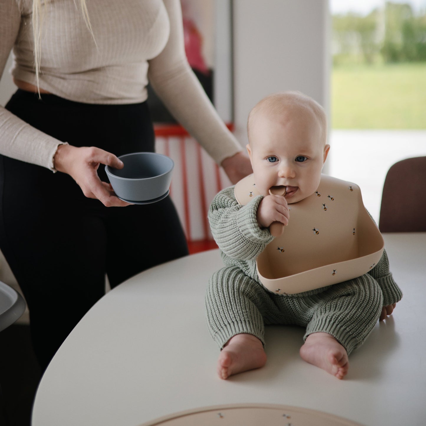 Baby sitting on a table wearing a bib and green outfit, with an adult holding a bowl in the background.