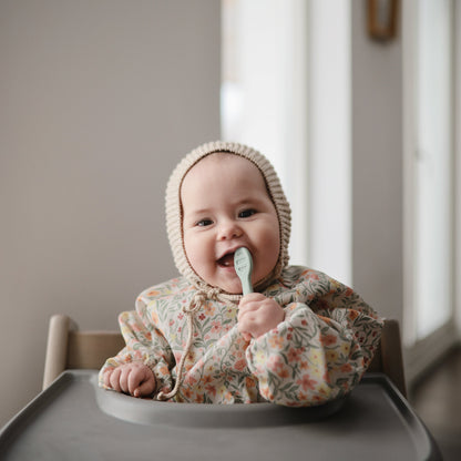 Baby in a floral outfit with a hood sitting in a high chair, eating with a spoon.