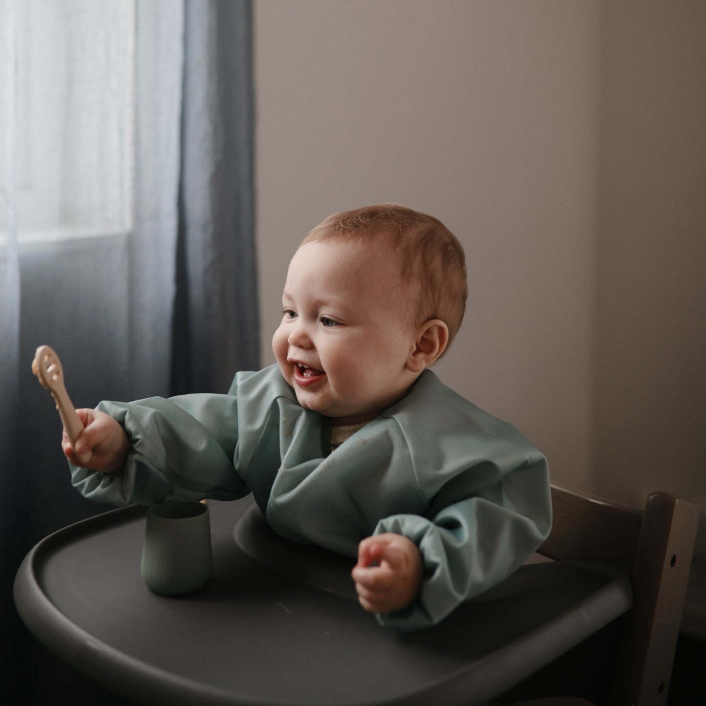 Baby in a high chair holding a wooden spoon, wearing a green bib.