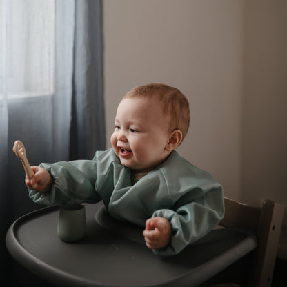 Baby in a high chair holding a wooden spoon, wearing a green bib.