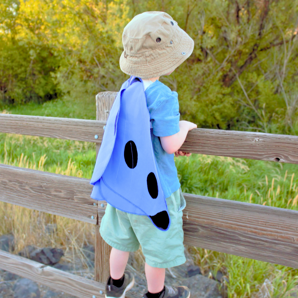Child wearing a blue dinosaur costume standing on a wooden fence with greenery in the background