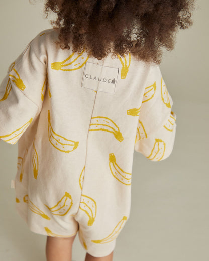 Child wearing a white outfit with yellow banana patterns and 'Claude' branding.