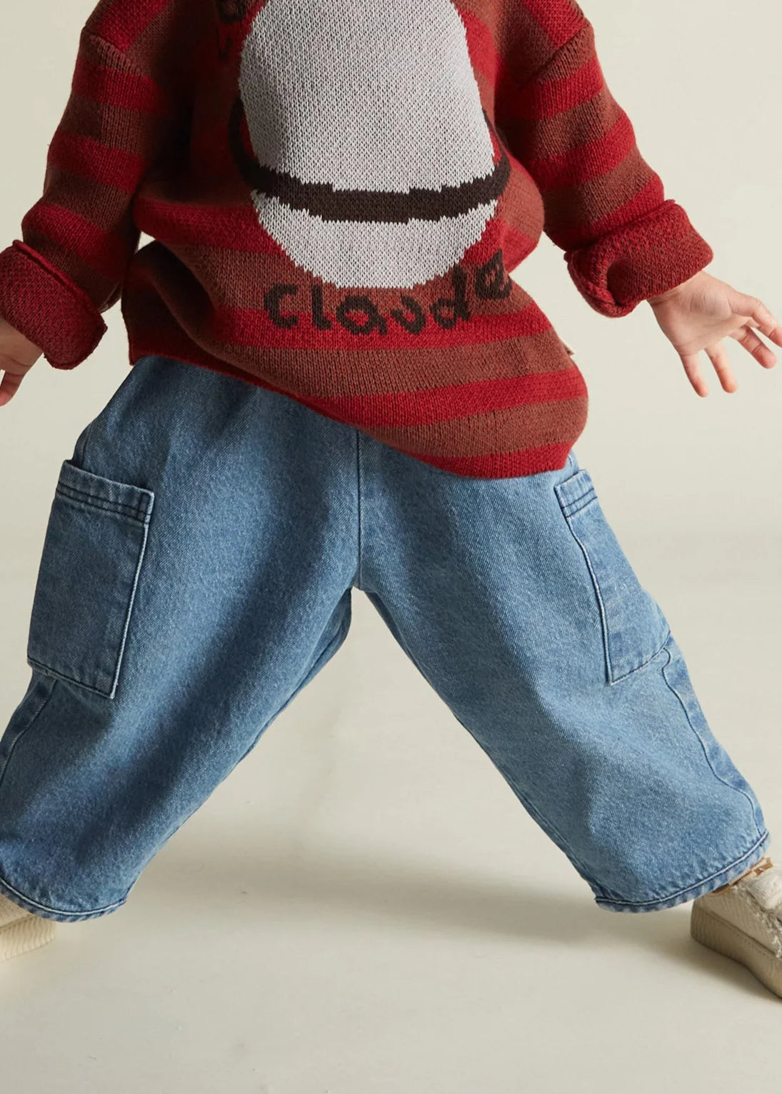 Child wearing a red and brown striped sweater with a graphic design and blue jeans on a beige background