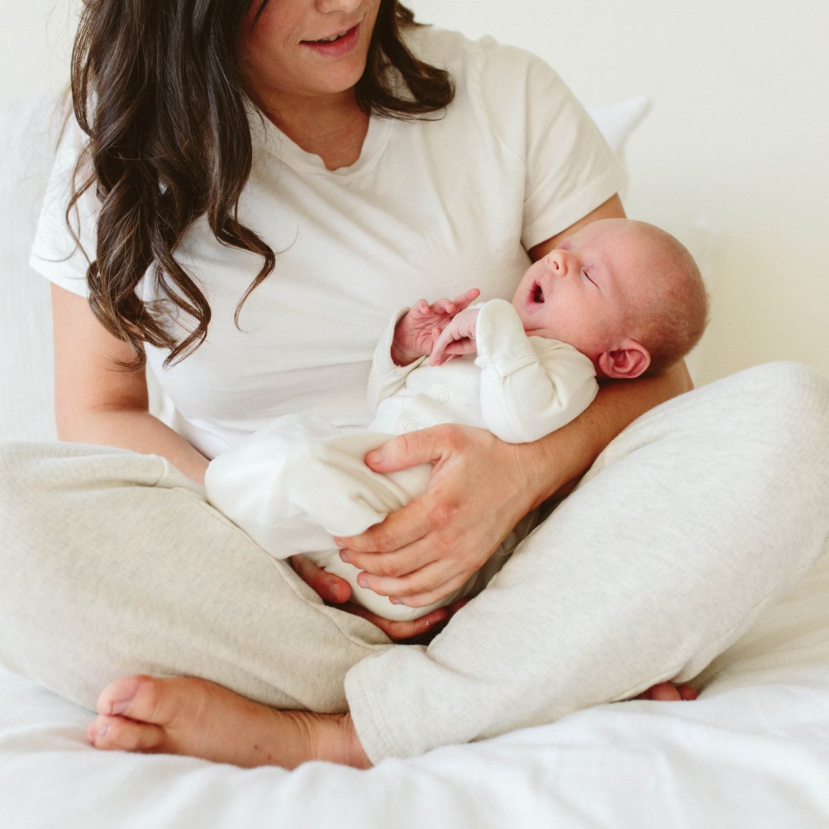 Woman holding a newborn baby wrapped in a white blanket on a white bed.