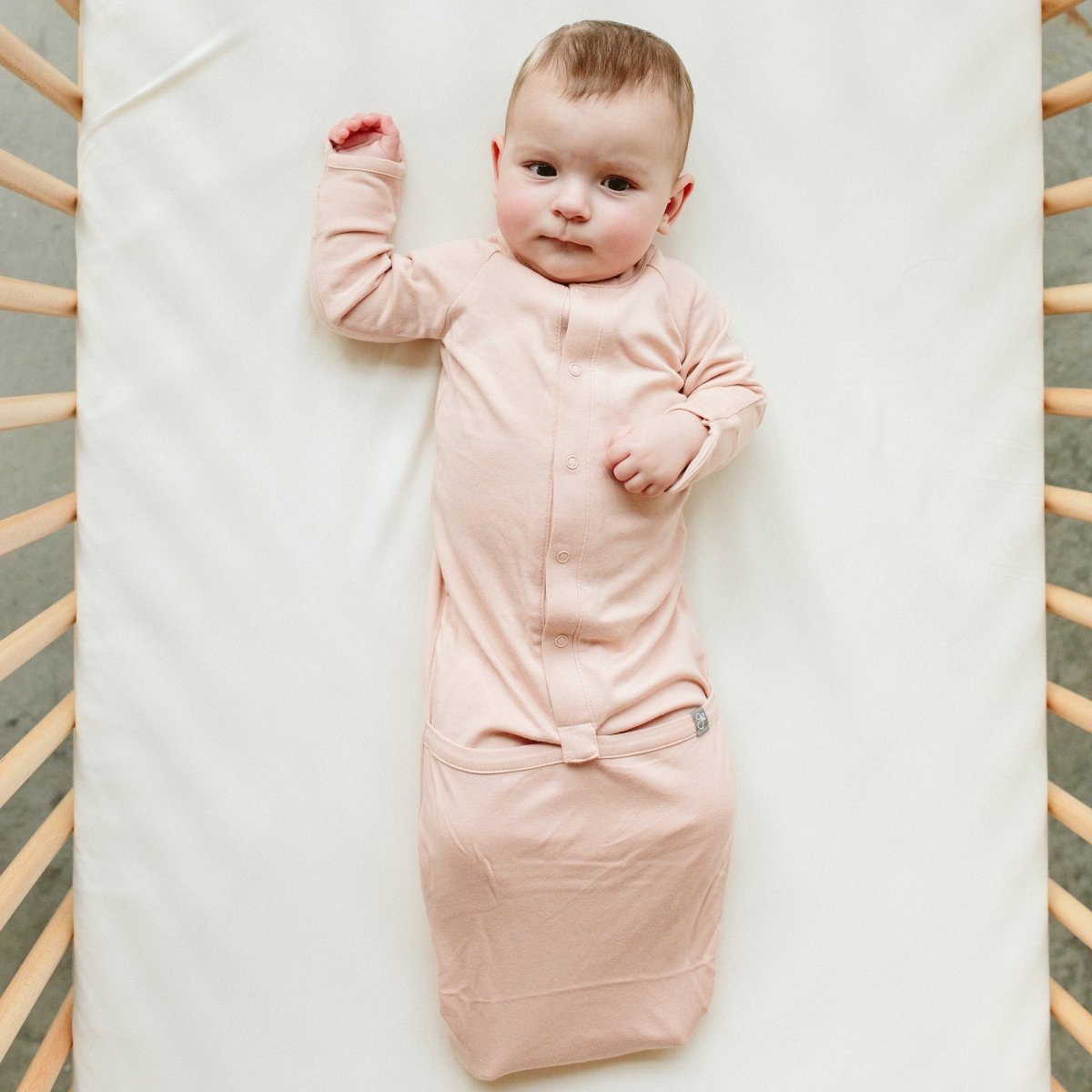 Baby in a pink sleep bag lying on a white blanket with a wooden crib frame.