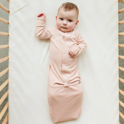 Baby in a pink sleep bag lying on a white blanket with a wooden crib frame.