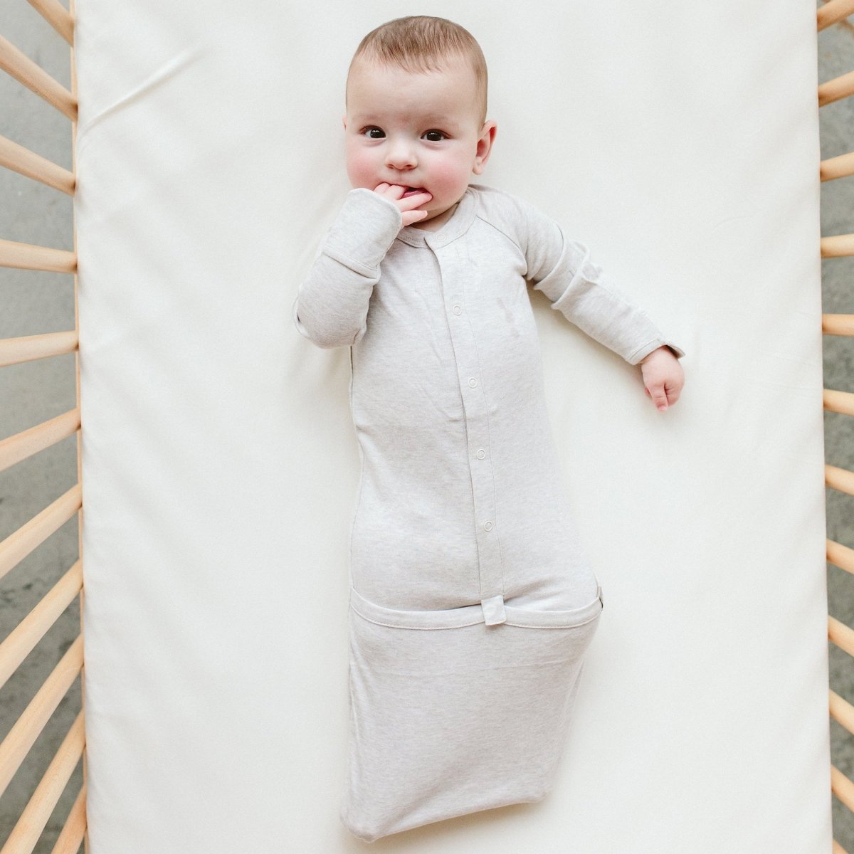 Baby in a light gray sleep bag lying on a white blanket with a wooden crib frame.