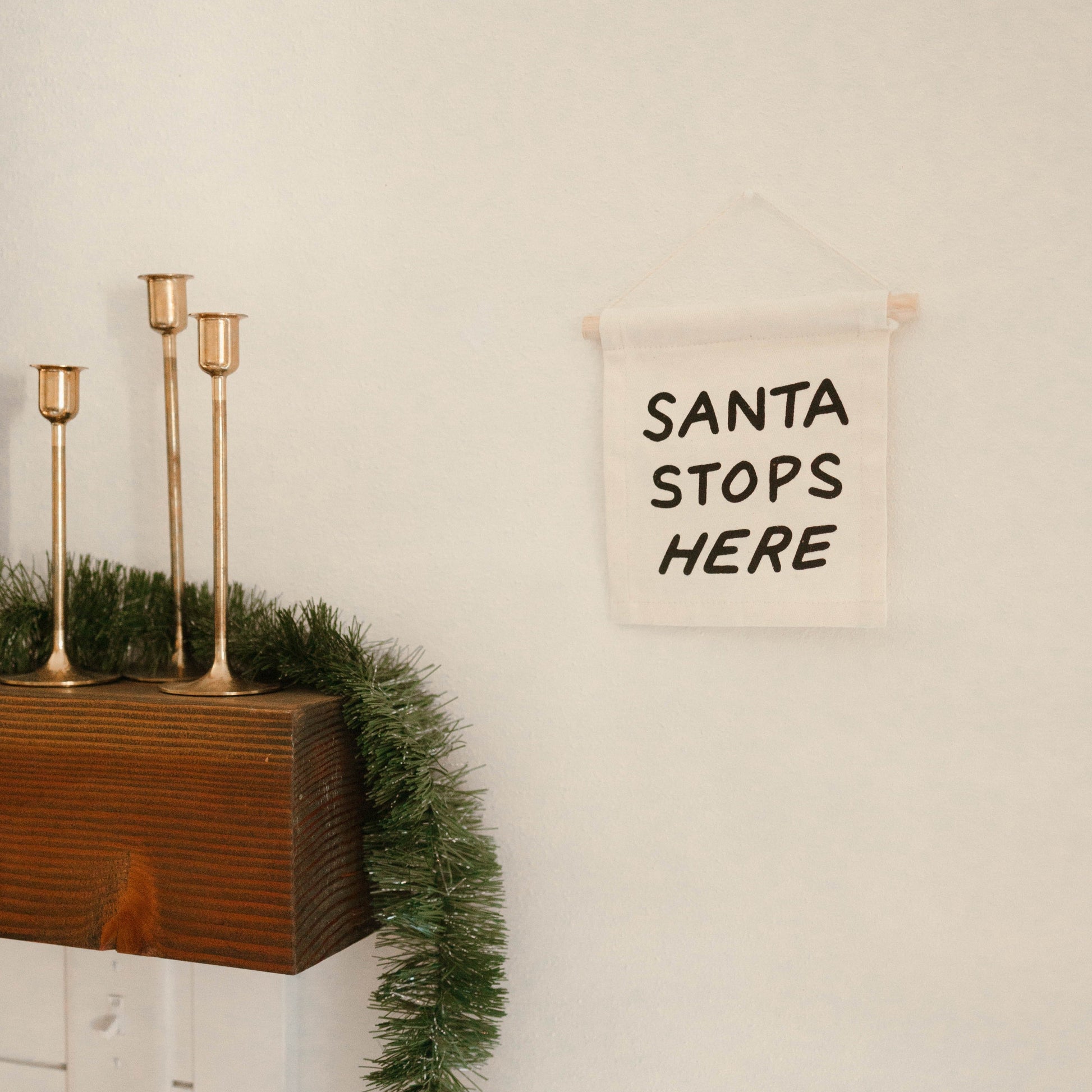 Decorative sign with 'Santa Stops Here' text on a wall above a wooden shelf with candles and greenery.