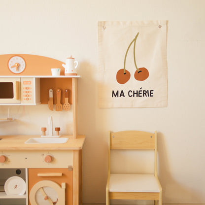 Children's play kitchen set with a chair and decorative wall hanging on a light beige wall.