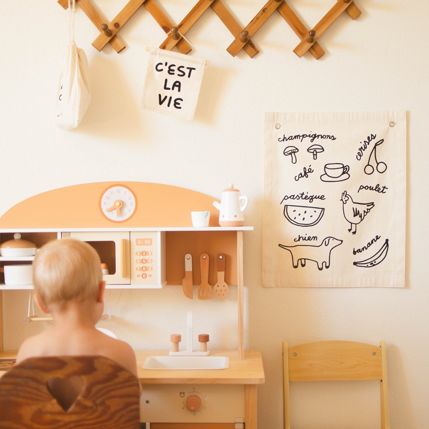 Child playing with a wooden play kitchen set in a room with a wall chart of French words and illustrations.