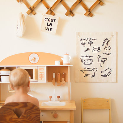 Child playing with a wooden play kitchen set in a room with a wall chart of French words and illustrations.
