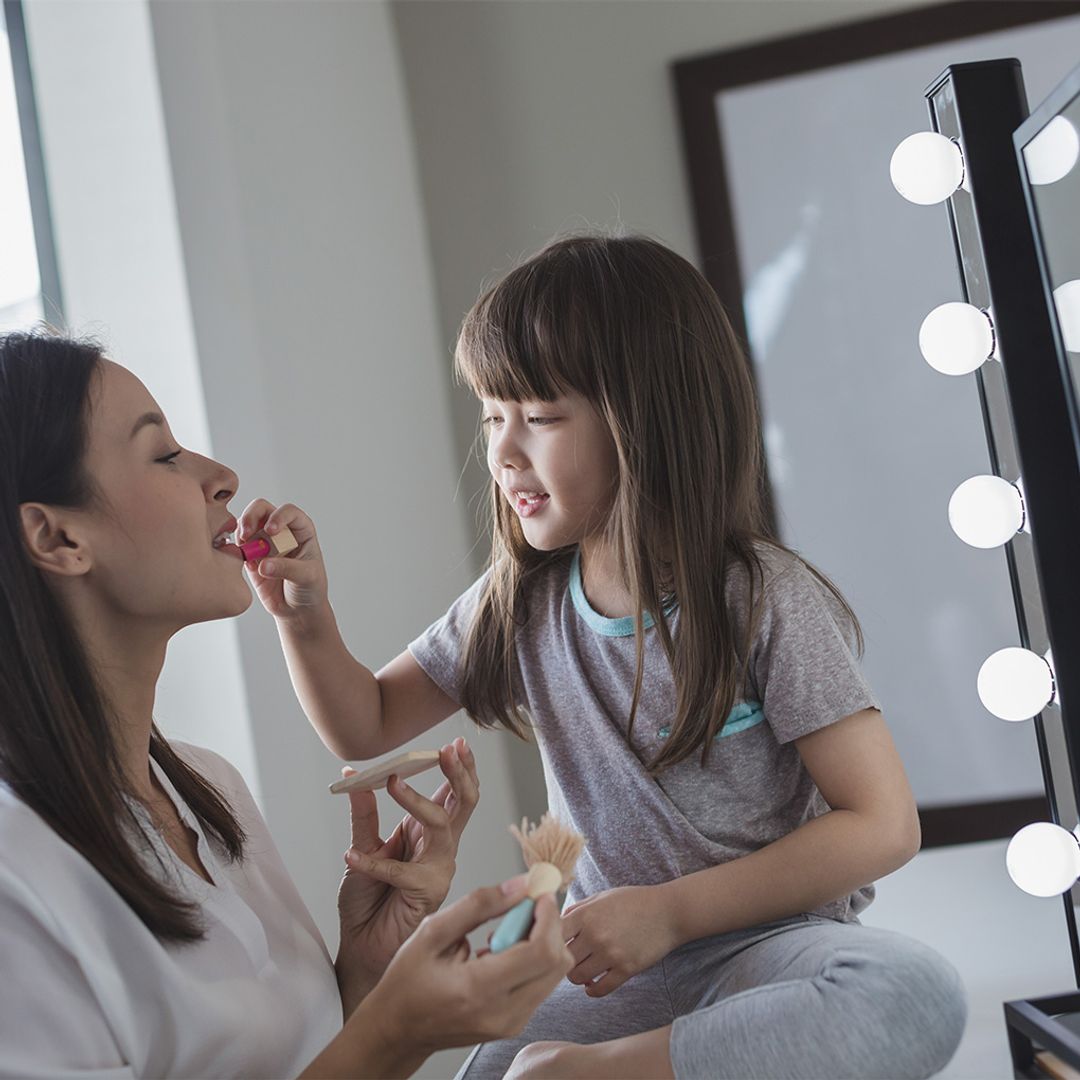 Woman and young girl playing with makeup in front of a mirror with light bulbs.